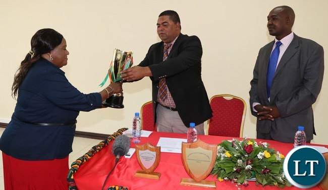 National Sports Council of Zambia Board Member Matilda Mwaba presents some independence tournament Trophies to Lusaka Province Minister Obvious Mwaliteta while his Deputy PS Japhen Mwakalombe looks on in Lusaka