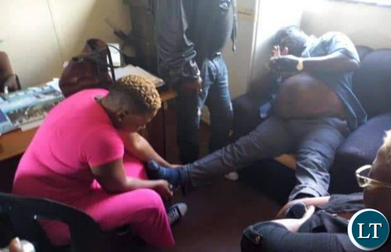 A tired looking Kambwili being held to remove his shoes at Woodlands Police Station by a family member.